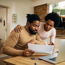 Un hombre y una mujer observan un papel juntos. Al frente de ellos hay una computadora portátil abierta en una mesa y un niño jugando atrás.