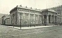 Un edificio de estilo neoegipcio en la esquina de una calle; hay columnas dóricas en la fachada a la derecha de la imagen. Hay hombres en la calle, y se está trabajando con grúas, en un edificio adyacente fuera de cuadro a la derecha.