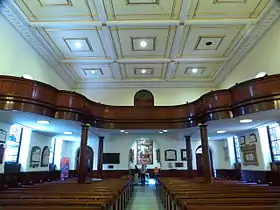 El interior siendo visto hacia una salida arqueada. Hay filas de bancas y un pasillo central. Hay un gran coro en madera obscura con una balastra curva a través de la parte trasera de la iglesia