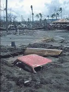 Rabaul luego de la erupción volcánica del 1994.                                  En la fotografía se aprecia un auto rojo tapado casi en su totalidad, de lo que parece ser ceniza y barro, apenas viéndose el techo, al fondo se ven casas destruidas y palmeras de las que solo quedo el tronco.