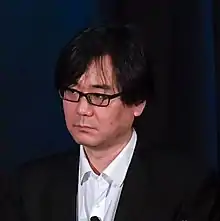 Un japonés de mediana edad con gafas delante de un micrófono.