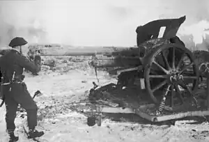 Fotografía en blanco y negro de Churchill uniformado mirando a través del cañón de un gran arma de artillería con una barricada de piedra visible detrás