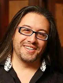 Fotografía en color del rostro de un hombre sonriente con largo cabello negro y gafas