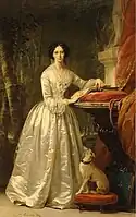 Retrato de mujer de pie y de cuerpo entero, con el pelo recogido y un vestido blanco sobre fondo oscuro neutro.