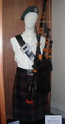 Fotografía del uniforme de Millin expuesto sobre un maniquí en una vitrina de museo, consistente en un gorro negro, una camisa blanca, un cinturón negro ancho con una gran hebilla de plata que cruza el pecho desde el hombro derecho hasta el lado izquierdo de la cintura, una gaita y una falda escocesa oscura de tartán multicolor
