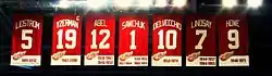 The banners of seven retired numbers. The banners, from left to right, read "Lidstrom 5" "Yzerman 19" "Sawchuk 1" "Delvecchio 10" "Lindsay 7" "Abel 12" "Howe 9". The Yzerman banner has a small "C" at the top right corner.
