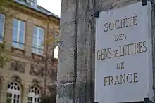 Photographie en couleur d'une plaque sur un pan de mur, blanche avec des caractères dorés