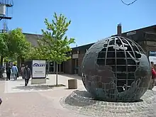 Gente caminando afuera de un edificio de ladrillo de una sola planta con escultura de globo metálico y letrero de biblioteca