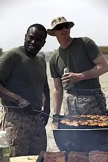 Dos varones en camisetas de mangas cortas idénticas y pantalones de camuflaje, uno de piel muy oscura sin sombrero y uno de piel muy clara con sombrero y anteojos de sol, parados sonriendo por sobre una barbacoa llena de carne cocida en una locación brillante